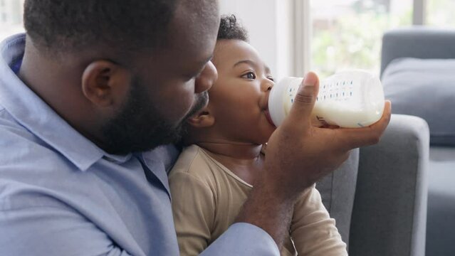 African father Sitting on sofa, holding and feeding newborn baby from bottle milk in living room. Authentic lifestyle candid real moment. Single dad family home life concept.