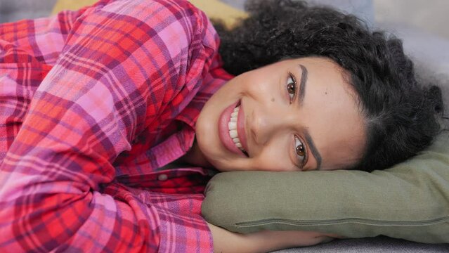 Close up of peaceful dark-haired lady relaxing with closed eyes on pillow indoors. Beautiful african american woman wearing bright checkered shirt opening eyes and smiling at camera.