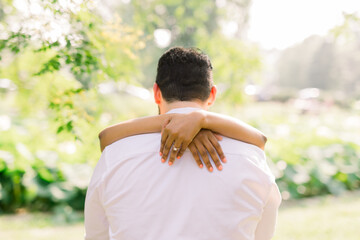 A South Asian dark skinned woman has her arms wrapped around her Caucasian fiance. She wears an engagement ring. 