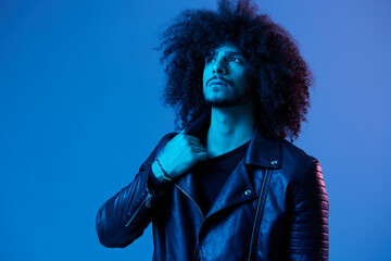 Portrait of fashion man with curly hair on blue background multinational, colored light, black leather jacket trend, modern concept.