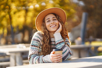 Portrait of a cute woman in a hat sits with a cup of coffee overlooking the lake. Outdoor photo of relaxed curly woman enjoying hot drink in autumn park.