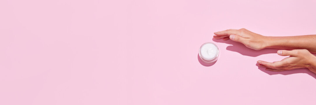 Banner. Top view of young tender woman's hands holding open jars of cosmetic creams on pink background. Female face care.