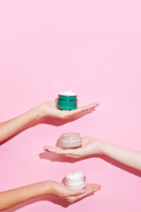 Open jars of cosmetic creams with womans hands on pink background. Female face care. Anti-aging. Treatments.