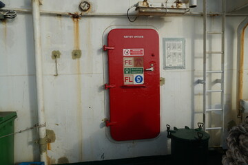 Red metal door with stickers is closing the entrance to safety locker on the container vessel. The wall in aft station of the ship are partly rusted has white colour.
