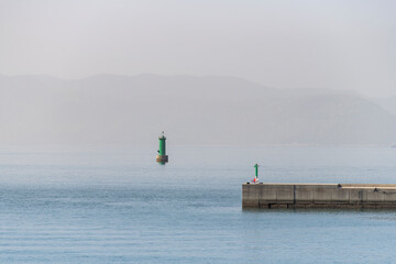 Partial view of the concrete wave barrier of Miyanoura Port on Naoshima Island, Japan with a tranquil sea, green small lighthouse in the water and background mountains in a haze of Kojin Island