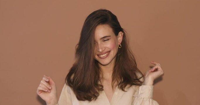 Upbeat brunette haired woman dances carefree has fun wears beige shirt, gold earrings isolated over beige background. Beautiful female model foolish around. Music, dance concept.