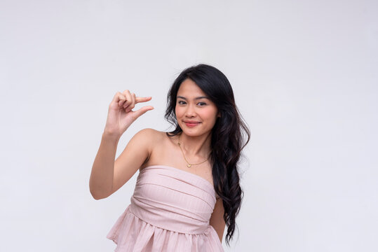 A polite young asian woman asking for a little bit more, gesturing with thumb and index finger. Isolated on a white background.