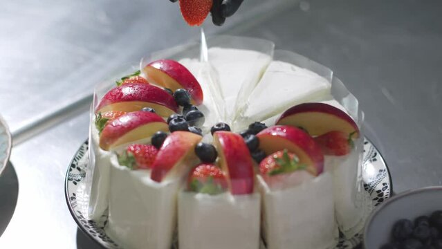 Close up of a chef's hand wearing black gloves laying an apple slices, blueberry, and strawberry slices in a circle on top of a cake. Decorate the cake.