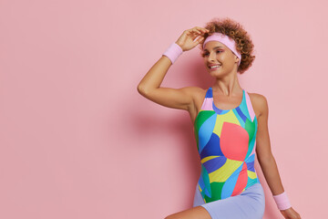 Cheerful woman with headband on curly haircut wearing colourful leotard doing exercises inside with smile, sport lifestyle concept, copy space.