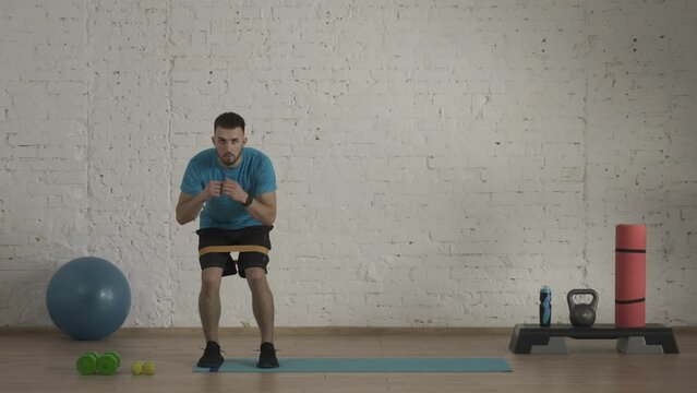 Man fitness instructor in sportswear doing half bend side steps with rubber band exercise for online class at the home studio. HDR BT2020 HLG Material.