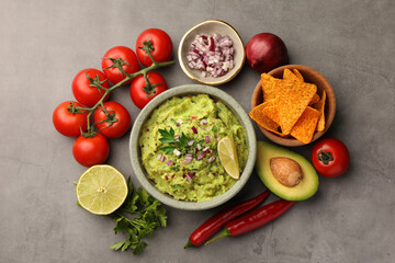 Bowl of delicious guacamole, nachos chips and ingredients on grey table, flat lay
