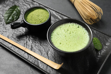 Cup of fresh matcha tea, bamboo whisk, spoon and green powder on black table, closeup