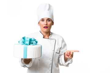Middle-aged chef caucasian woman with a big cake over isolated background surprised and pointing side