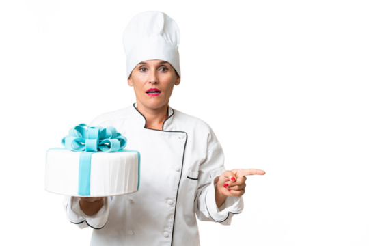 Middle-aged chef caucasian woman with a big cake over isolated background surprised and pointing side