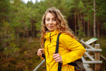 Female tourist in a yellow coat with a backpack traveling along a wooden path in the forest....