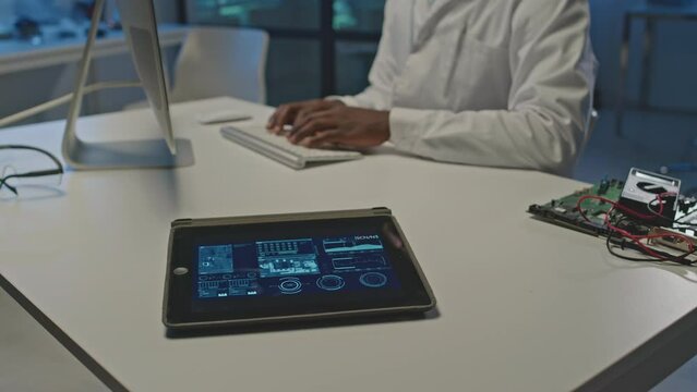 Selective focus on digital tablet screen with scheme of microchip at white desk in modern laboratory with hands of unrecognizable electronics engineer typing on computer keyboard in blurred background