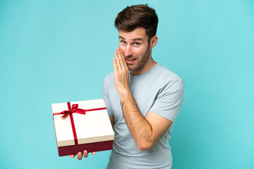 Young caucasian man holding a gift isolated on blue background whispering something