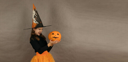 Close-up portrait of a funny teenage girl in a witch costume on a gray background, holding pumpkins in her hands. Banner