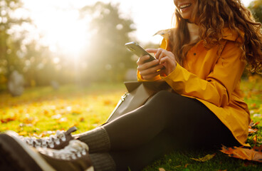 Cheerful woman with a smartphone in her hands sits in a clearing among yellow fallen leaves in an autumn park. Woman in a yellow coat spends time on an autumn weekend.