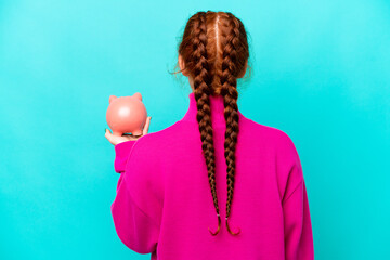 Young caucasian reddish woman holding a piggybank isolated on blue background in back position