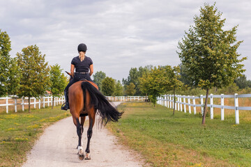Female rider, horseback riding along the trail that leads between white wood fences and fields. Equestrian leisure activity concept. - Powered by Adobe