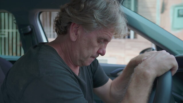Old man experiencing sadness holding on into car's steering wheel parked on street sidewalk. Desperate senior person suffering from depression struggling with daily activities