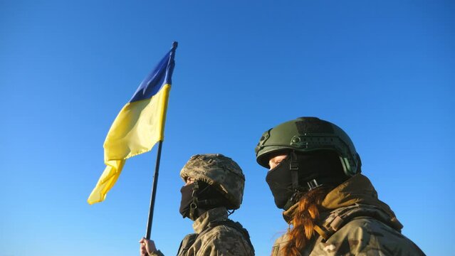Young soldiers of ukrainian army standing at peak of hill with raised flag of Ukraine. Military couple in camouflage uniform taking each other hands as a symbol of support. Victory at war. Crane shot