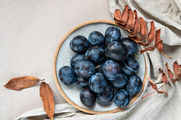 Ripe plum fruits on plate on neutral beige table and linen tablecloth background with dried fall leaves