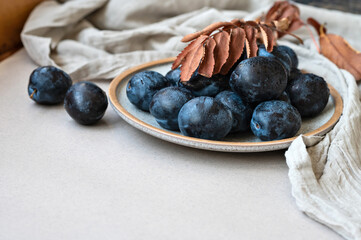 Fresh aesthetic plum fruits on plate, linen tablecloth, dried fall brown leaves on neutral beige background