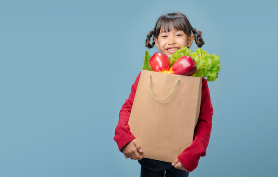 A little girl is holding a paper bag filled with fruits and vegetables.