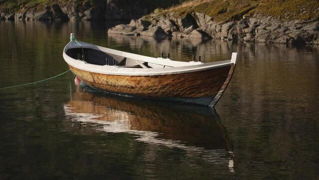 Against the backdrop of towering mountains, a wooden rowing boat finds repose on a buoy in a Norwegian fjord, its reflection on the serene waters adding an ethereal touch to the scene