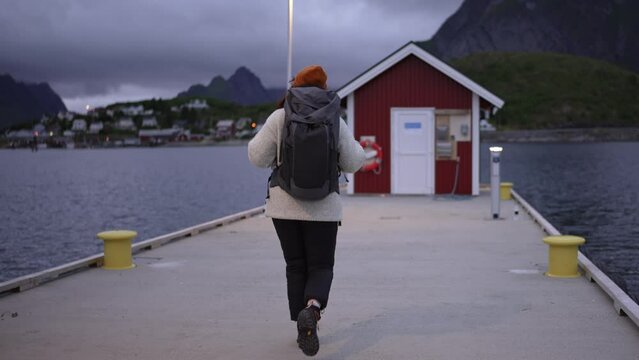A young girl, donning a hat, backpack, and wool sweater, walks towards the pier on the Lofoten Islands in Norway, as seen from behind, embodying the essence of a traveler's adventure