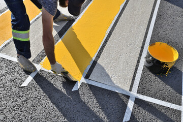 A worker applies yellow paint with spatula to the asphalt at a pedestrian crossing.