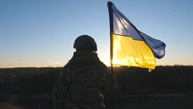 Ukrainian army woman stands to the peak of hill with waving blue-yellow banner at sunset. Young female soldier in camouflage uniform lifting flag of Ukraine. Resistance to russian invasion concept