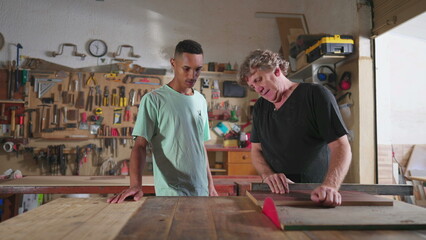 Carpenter and apprentice working at workshop, teacher orienting young man with saw machine at...