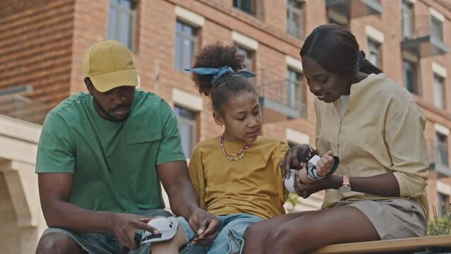 Medium long shot of African American parents helping little girl to put on protective wrist guards and knee pads for roller skating outdoorsMedium long shot of African American parents helping little 
