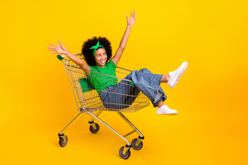 Full length photo of cheerful funky nice girl sit inside market trolley raise arms look empty space isolated on yellow color background - obrazy, fototapety, plakaty