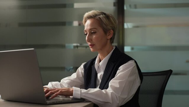 Confident middle-aged businesswoman female CEO worker in suit sitting at desk working typing on laptop computer looking to screen with serious expression, in corporate office. Shooting in slow motion.