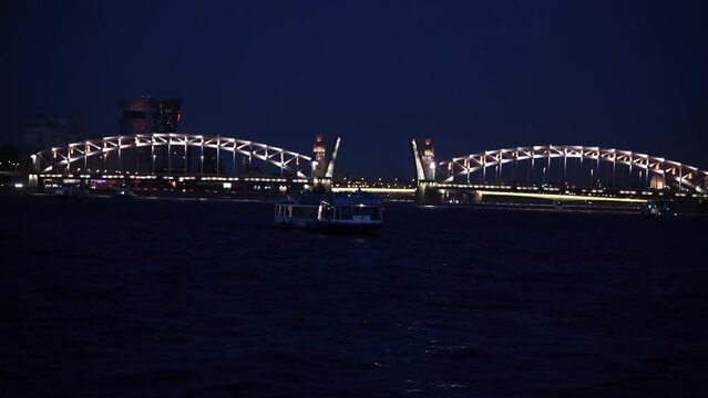 Tour boats sails next to open illuminated Bolsheokhtinsky bascule bridge at night. Real time handheld video. Soft focus. Water tourism theme.