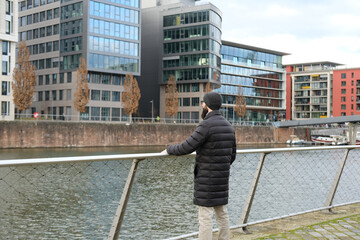 young bearded man in coat and hat stands on shore river Main, winter walks in Frankfurt, View of modern residential buildings, Gutleitviertel district, boats and yachts moored on banks