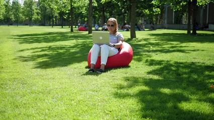 A young woman with a laptop is sitting in a red upholstered chair in a modern park, she closes her laptop and looks at the camera. A female freelancer in sunglasses works in a park. Stable image.