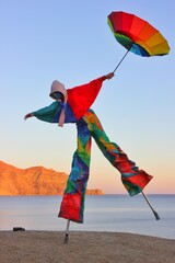 A man on stilts in a carnival costume stands with a rainbow umbrella on one leg on the seashore against a blue sky 