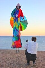 A model in a multi-colored suit on stilts and a mask poses for a photographer on the seashore....
