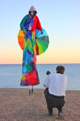 A model in a multi-colored suit on stilts and a mask poses for a photographer on the seashore. Colored costume for participation in a carnival or celebration. umbrella of all colors of the rainbow