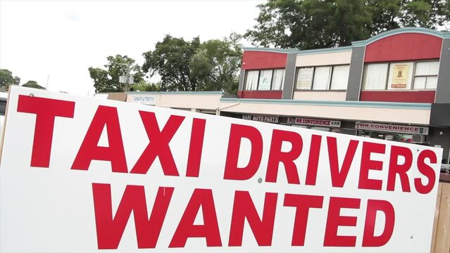 taxi drivers wanted sign in red capital letters writing text with buildings and trees behind