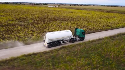 Aerial 4k drone tracking shot of Bulk Dry Cement trailer truck driving down a dusty rural road. Construction machinery off road. Cement powder transported to remote construction site among farms.
