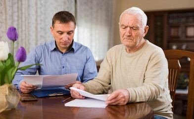 Senior man filling out papers with help of his adult son in living room at home