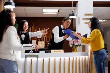 Hotel guest receives registration form from asian male receptionist to make room reservation. Smiling female concierge giving direction to customer at front desk of cozy resort