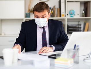 Worried businessman in protective mask working alone with laptop and papers in office. New normal business process during pandemic situation