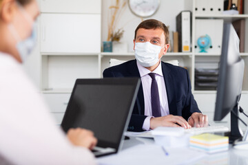 Portrait of busy entrepreneur in medical face mask sitting at office desk with papers and laptop. Concept of precautions and social distancing in coronavirus pandemic